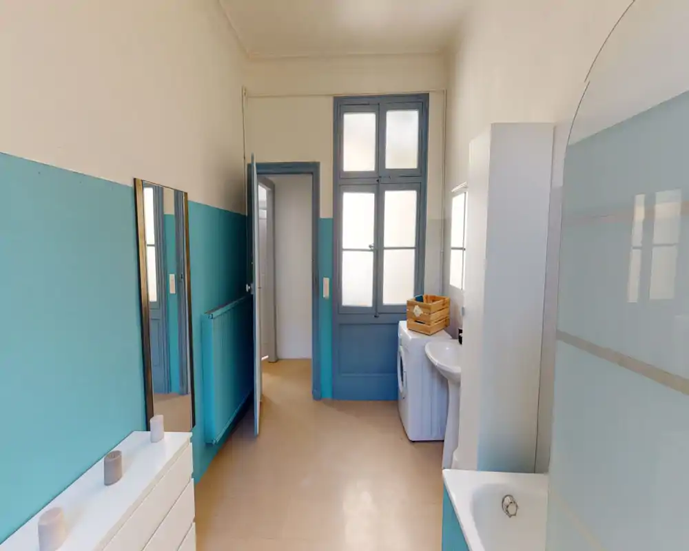 Salle de bains lumineuse et soignée avec baignoire, lavabo, machine à laver et rangement; murs bleu pâle et fenêtres dépoli pour intimité et lumière douce.