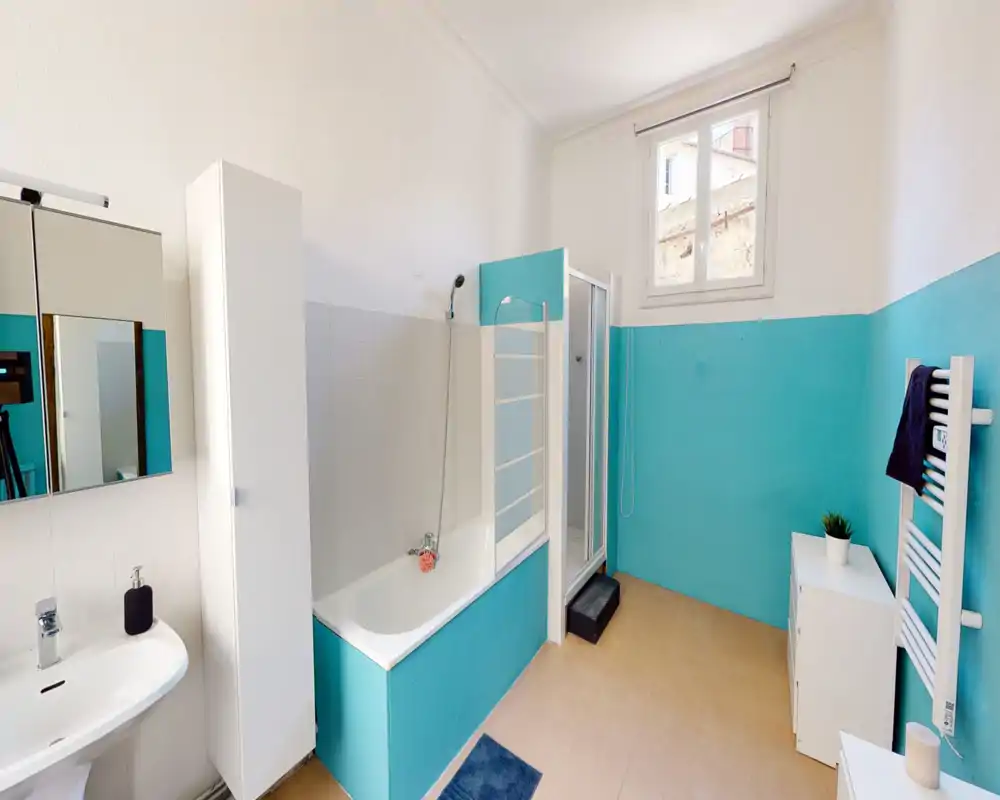 Salle de bain lumineuse et bien entretenue avec baignoire et cabine de douche séparée, carrelage turquoise mi-hauteur, lavabo mural et armoire de rangement — parfaite pour une annonce locative mettant en avant une salle d'eau complète.