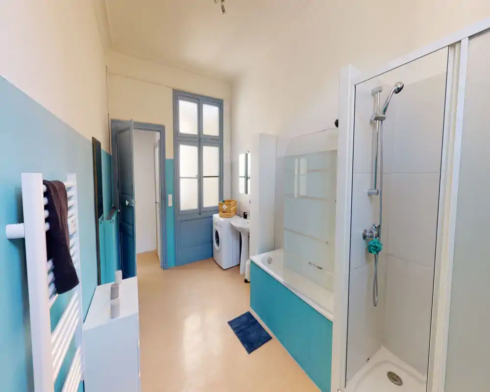 Salle de bains spacieuse et lumineuse avec cabine de douche séparée, baignoire avec paroi vitrée, lavabo et lave-linge — accents turquoise et grande fenêtre dépoli pour une atmosphère fraîche.