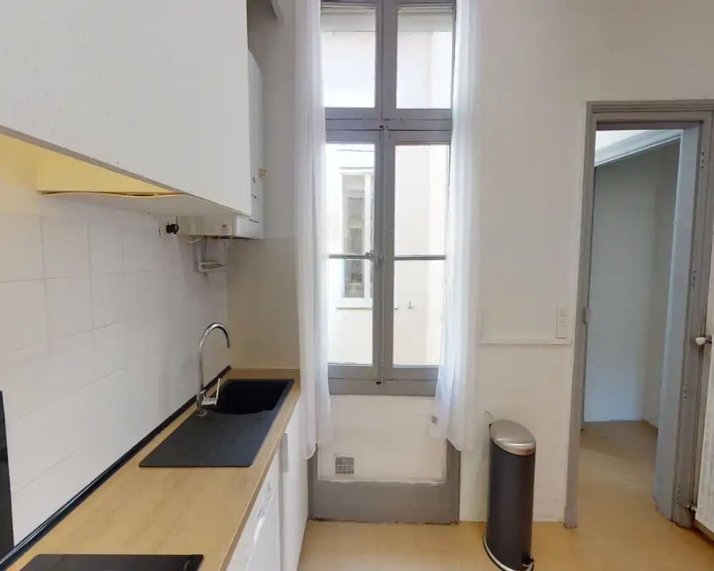 Cuisine compacte et lumineuse avec plan de travail en bois, évier intégré et plaque à induction; une grande fenêtre apporte la lumière naturelle et une disposition fonctionnelle adaptée à un petit appartement.