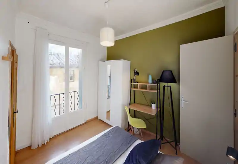 Chambre lumineuse et bien décorée avec un lit au premier plan, une armoire blanche et un petit bureau devant un mur vert — grande fenêtre apportant beaucoup de lumière naturelle.