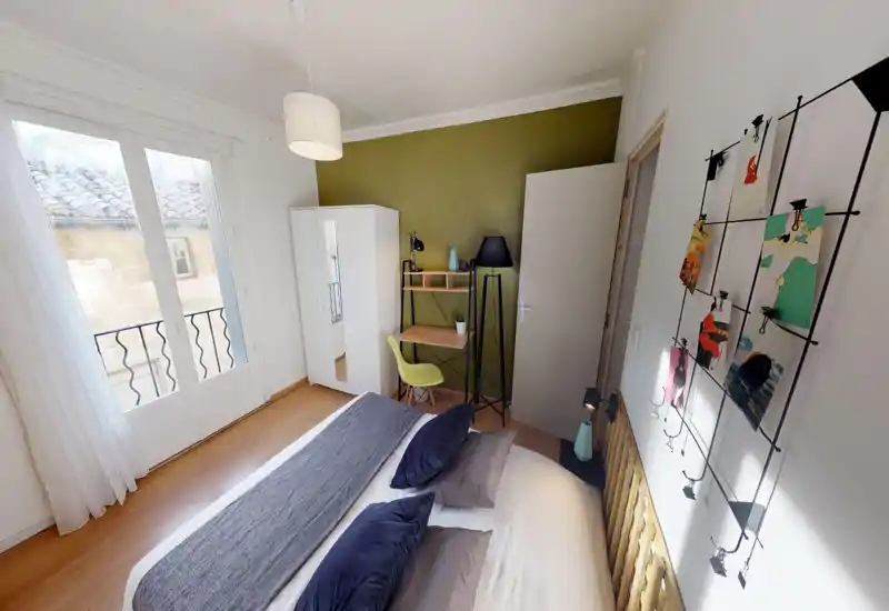 Chambre lumineuse et soignée avec un grand lit au premier plan, lumière naturelle via des portes-fenêtres, petit bureau et armoire contre un mur vert olive — idéale pour un séjour confortable en solo ou en couple.