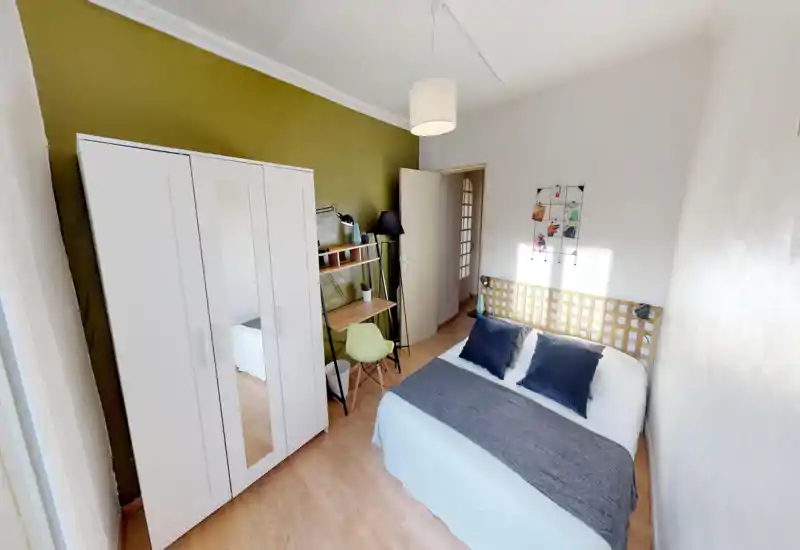 Chambre lumineuse et soignée avec lit double, textiles confortables et une armoire. La lumière naturelle met en valeur la tête de lit en bois et un petit espace de travail, idéale pour les séjours.