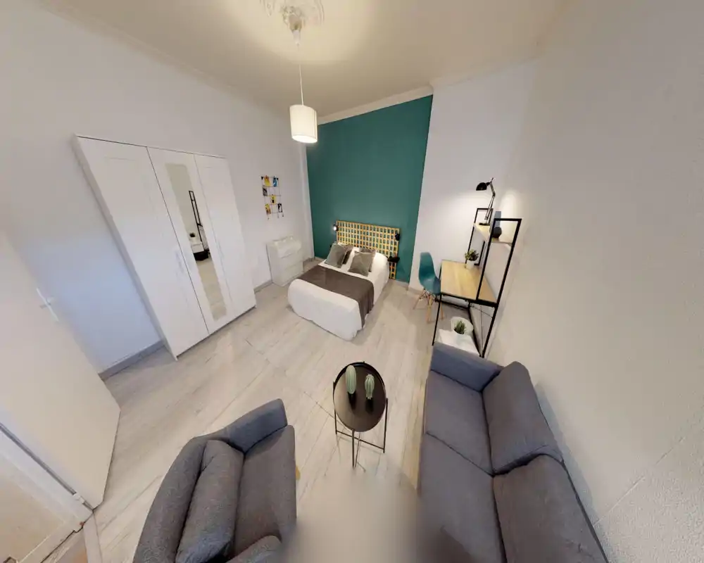 Bright, well-styled bedroom with a double bed against a teal accent wall, wardrobe, desk and a small seating area — ideal for a cozy studio stay.