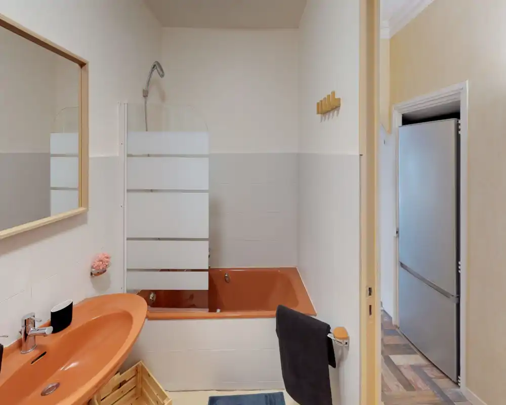 Salle de bain compacte avec lavabo orange et baignoire assortie, paroi de douche en verre, miroir et porte-serviette — lumineuse et bien détaillée.