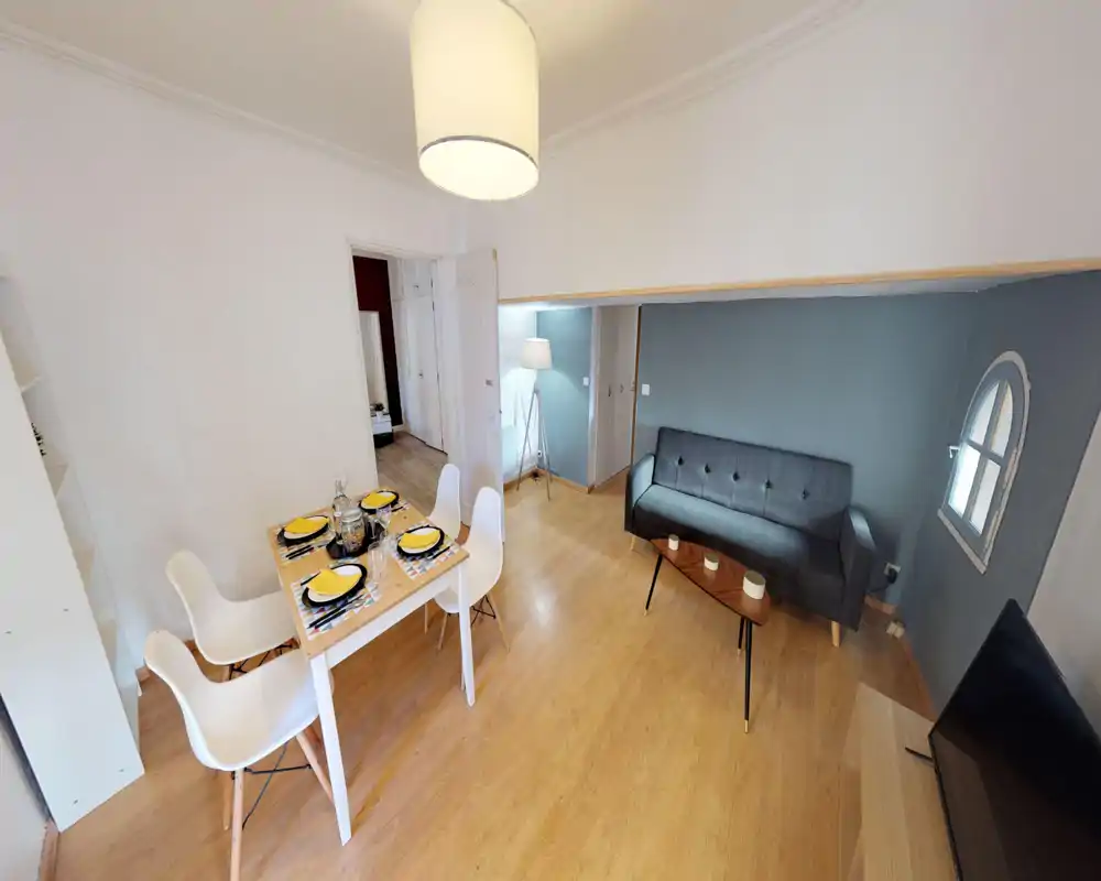 Espace séjour lumineux et soigné avec un canapé gris moderne, une table basse et une petite table à manger pour quatre. Sol en bois, éclairage élégant et un mur d'accent bleu apaisant.