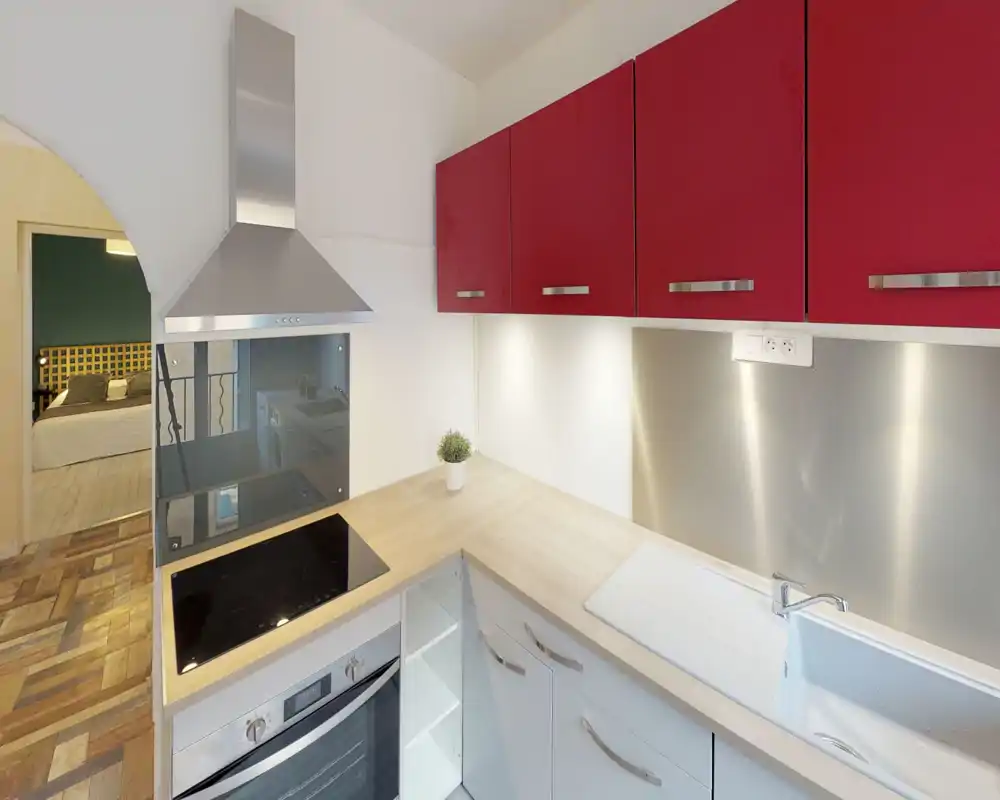 Cuisine compacte et moderne avec plaque de cuisson électrique, hotte en inox, meubles hauts rouges et plan de travail en bois clair; évier intégré et éclairage sous les placards pour un espace de travail lumineux et fonctionnel.