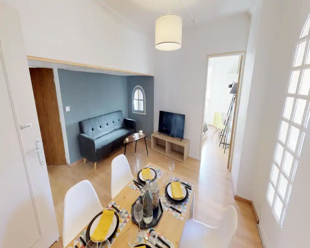 Espace séjour/salle à manger lumineux et moderne avec un petit canapé, table basse et TV, ainsi qu'une table dressée au premier plan — espace aéré et bien mis en scène.