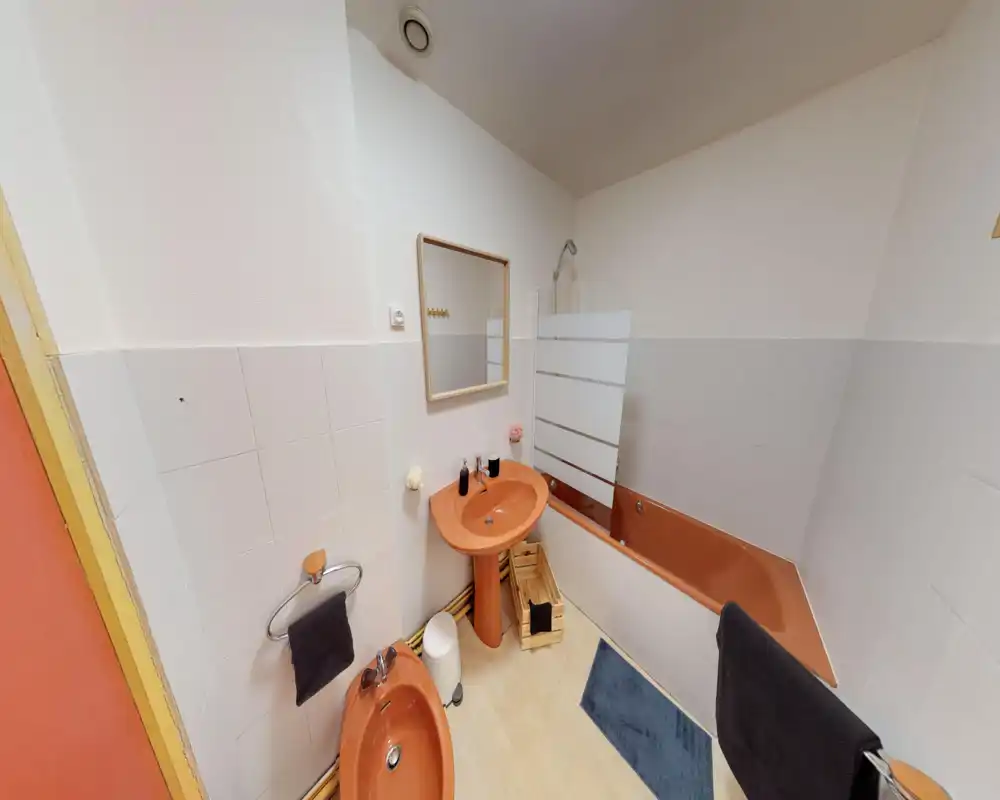 Salle de bains propre et lumineuse avec lavabo, bidet et baignoire orange assortis, murs carrelés blancs, miroir et pare-douche — nette et prête à accueillir des invités.