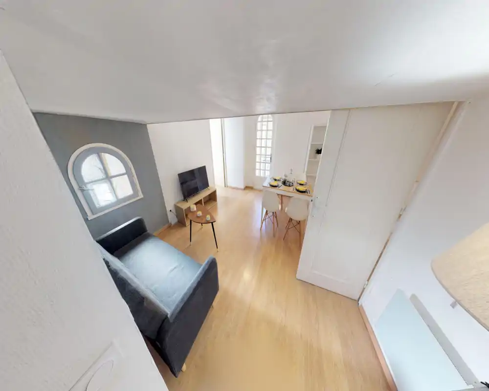 Petit salon lumineux avec un canapé sombre deux places, une table basse, une TV sur meuble bas et un coin repas pour quatre personnes — moderne et bien éclairé.