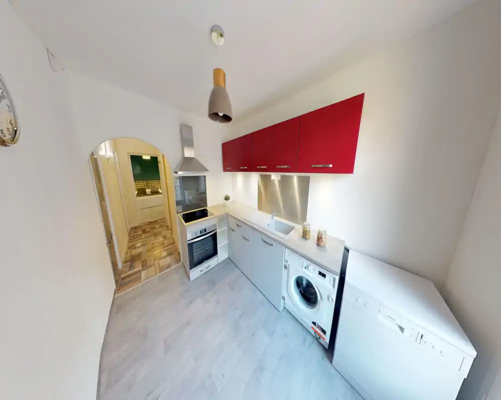 Cuisine moderne et compacte avec éléments hauts rouges, four et hotte intégrés, évier, lave-linge et lave-vaisselle. Surfaces propres et lumineuses, plan de travail en L pratique pour un petit appartement.