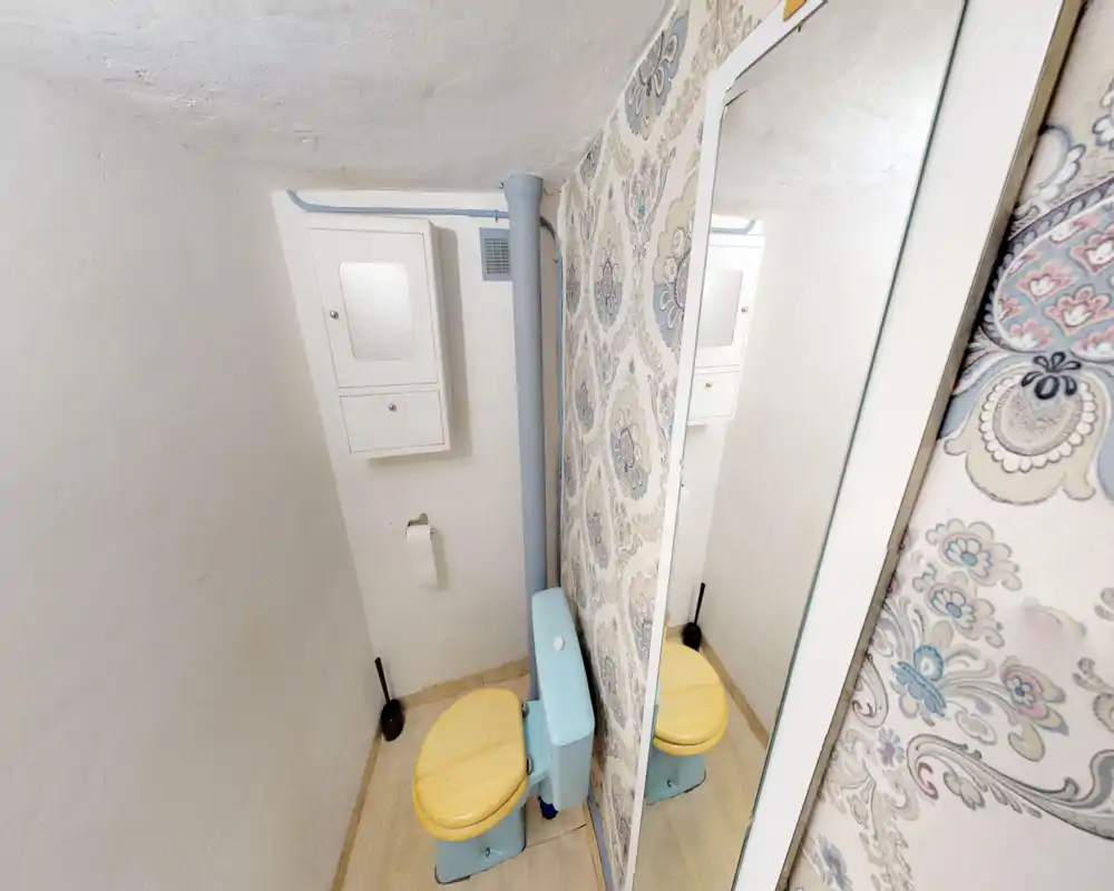 Salle de bains compacte avec WC style vintage pastel, abattant en bois et papier peint à motif. L'espace est petit, propre et bien éclairé.