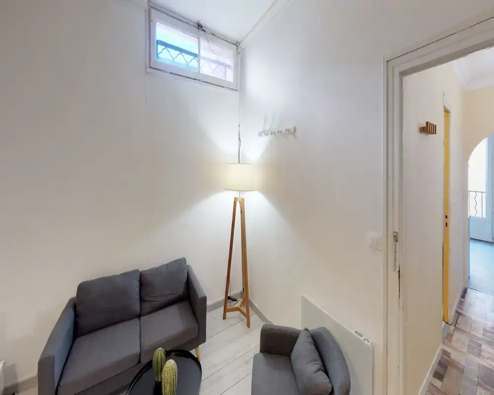 Petit salon bien mis en scène avec canapé et fauteuil gris, lampe sur trépied et table basse ronde — décoration lumineuse et neutre, idéale pour un espace locatif compact.