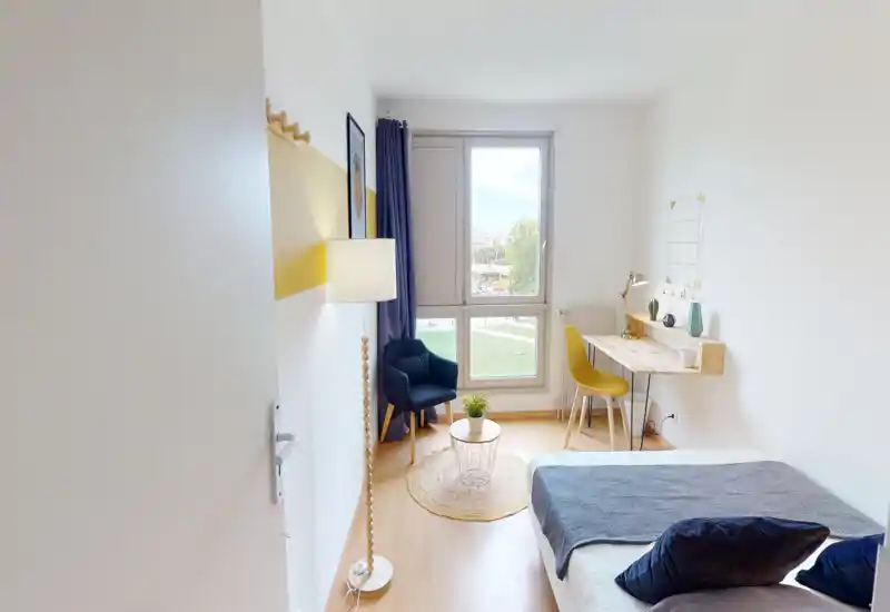 Chambre lumineuse et moderne avec un lit bien fait au premier plan, un fauteuil confortable près de la fenêtre et un bureau soigné — idéale pour travailler et se détendre.