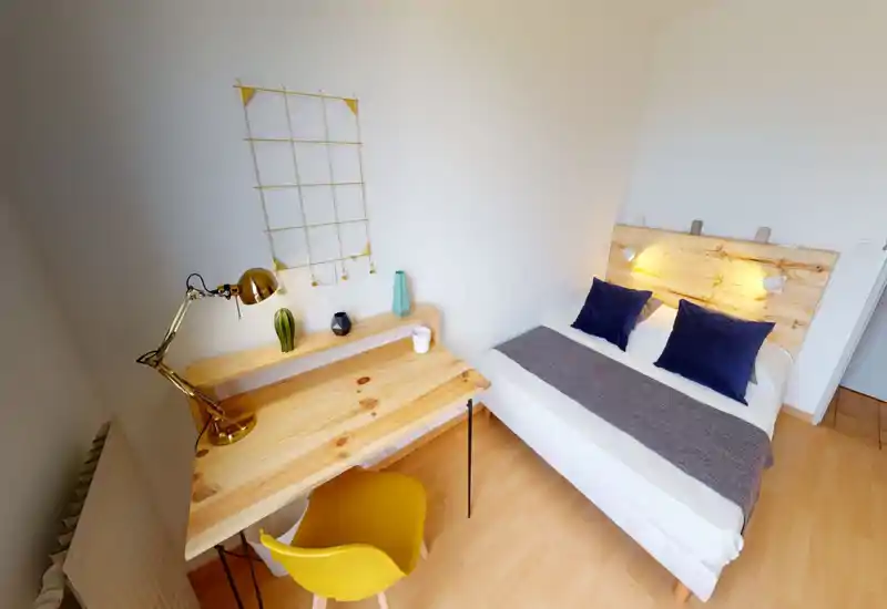 Chambre lumineuse et bien décorée avec tête de lit en bois, lit double avec coussins bleu marine et jeté texturé, et un petit bureau en bois avec chaise jaune — idéale pour un séjour cosy ou pour travailler.