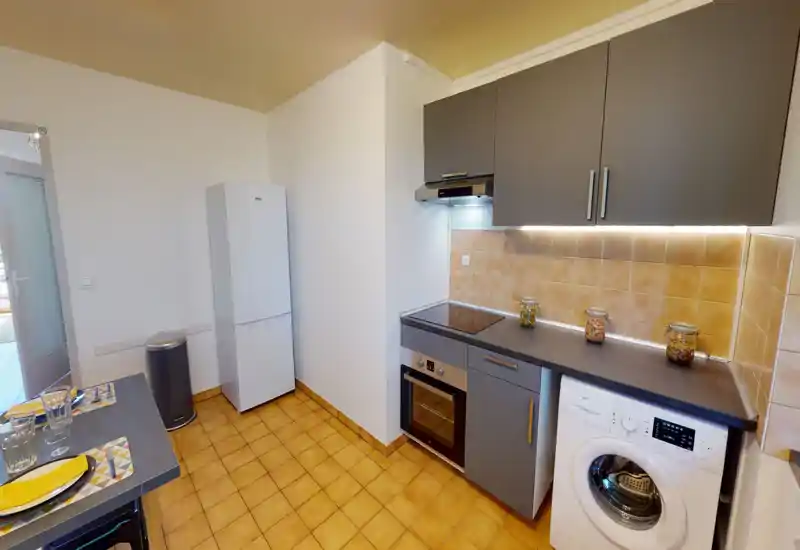 Cuisine compacte et moderne avec placards gris, four encastré, plaque à induction et lave-linge sous le plan de travail. Carrelage au sol lumineux et éclairage sous les meubles pour un espace de cuisson fonctionnel et soigné.
