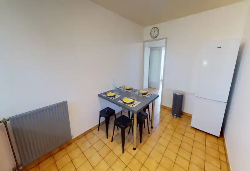 Coin repas simple et lumineux avec une table dressée pour quatre, tabourets métalliques et un réfrigérateur indépendant — espace fonctionnel et minimaliste pour repas décontractés.