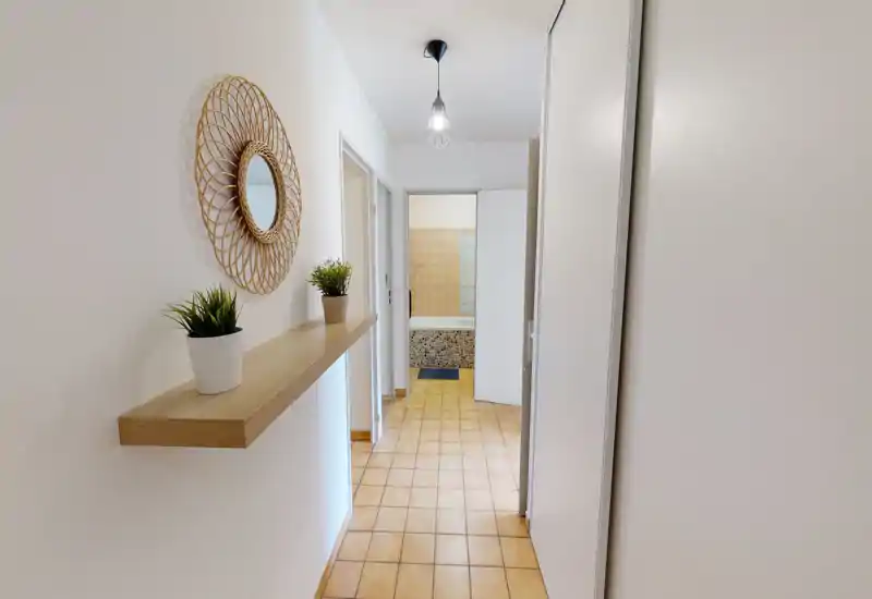 Entrée lumineuse et soignée avec une étagère murale en bois, un miroir décoratif et des plantes en pot. Le sol en carreaux terre cuite mène aux pièces adjacentes et à une salle de bain visible au fond.