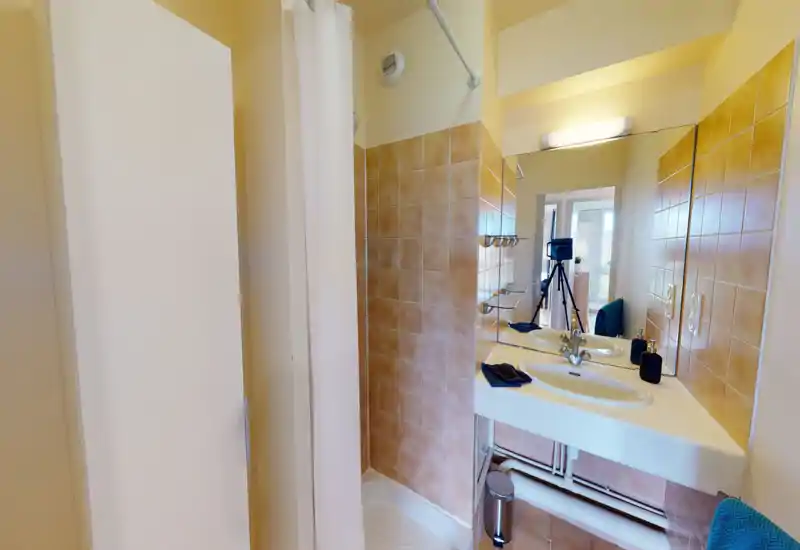 Salle de bains compacte et bien éclairée avec coin douche carrelé, lavabo mural visible dans le miroir et accessoires (serviette, distributeurs de savon).