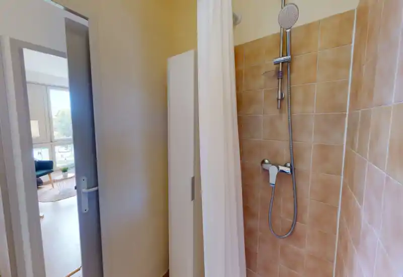 Douche carrelée compacte avec pommeau de douche chromé à main et carreaux beige neutre ; espace lumineux et propre avec un rideau de douche blanc.