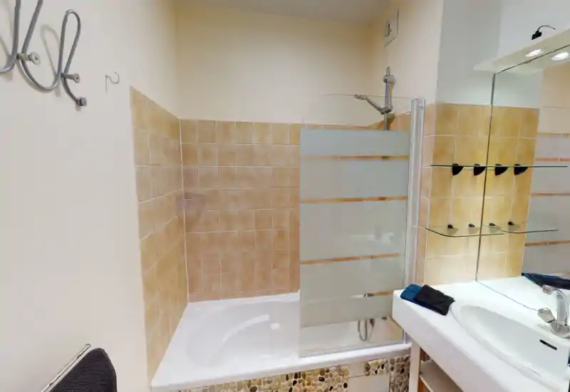 Salle de bains propre et bien éclairée avec baignoire/douche, murs carrelés, pare-douche en verre et lavabo avec miroir et étagères.