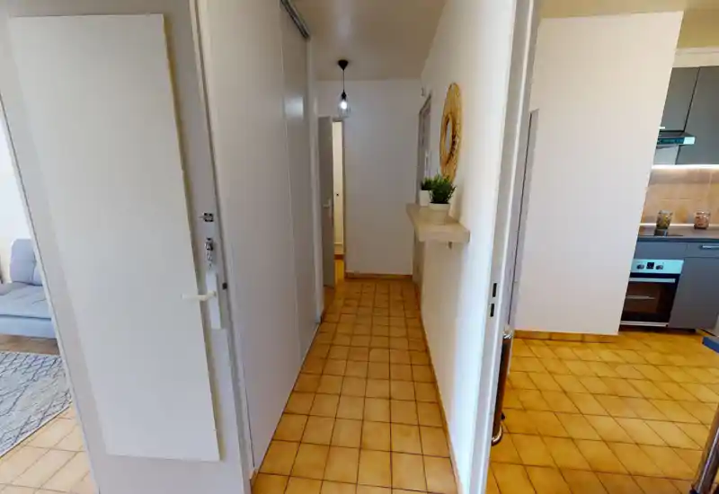 Entrée lumineuse et soignée avec sol carrelé, petite étagère avec plantes et miroir décoratif; portes vers la cuisine et le séjour.