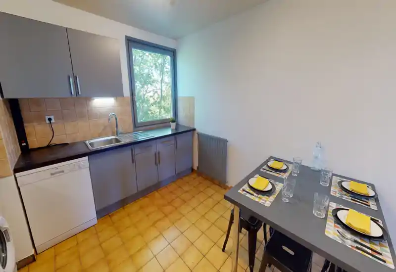 Cuisine compacte et soignée avec évier intégré et lave-vaisselle, fenêtre apportant de la lumière naturelle et petite table dressée pour quatre — idéale pour une cuisine locative fonctionnelle.