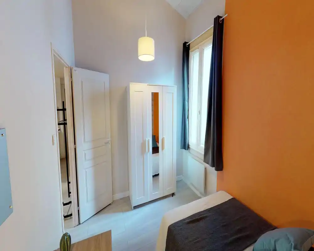 Compact bedroom with a double bed partially visible, a white wardrobe with mirrored panel, and a large window letting in natural light — warm orange accent wall adds character.