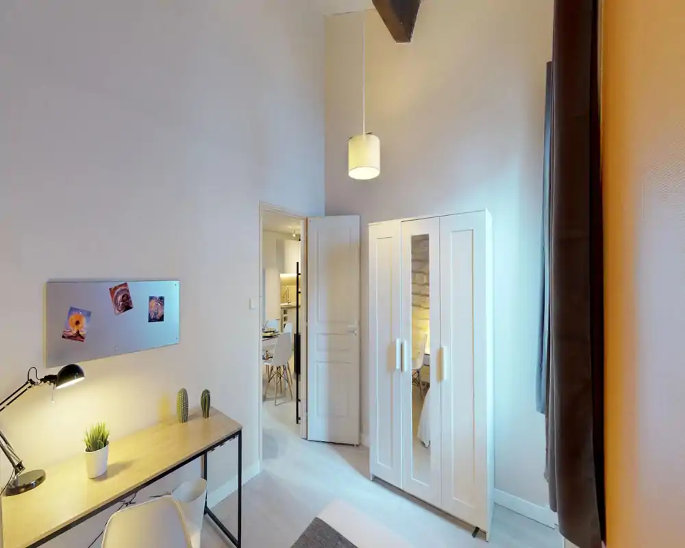 Bright, minimalist bedroom with a white wardrobe, a desk with lamp and plants, and a partially visible bed in the foreground. High ceiling and soft lighting create an airy, calm atmosphere ideal for short-term rentals.