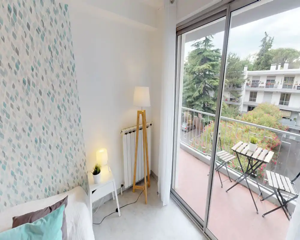 Coin de chambre lumineux et chaleureux avec lit partiellement visible, table de chevet et lampadaire, ouverture sur un balcon avec petite table bistrot et verdure.