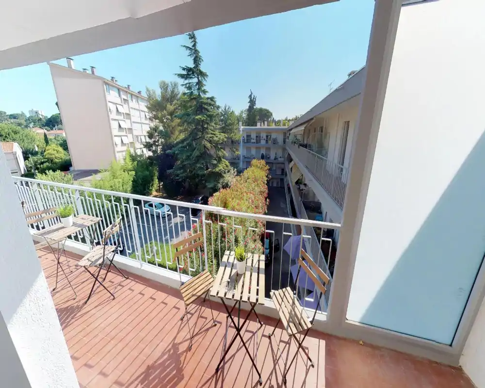Balcon ensoleillé avec petite table et chaises, donnant sur une cour arborée et des bâtiments voisins — idéal pour un café le matin ou se détendre à l'extérieur.