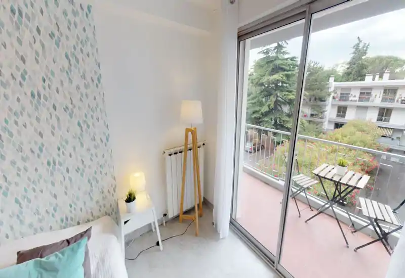 Coin de chambre lumineux et chaleureux avec lit partiellement visible, table de chevet et lampadaire, ouverture sur un balcon avec petite table bistrot et verdure.