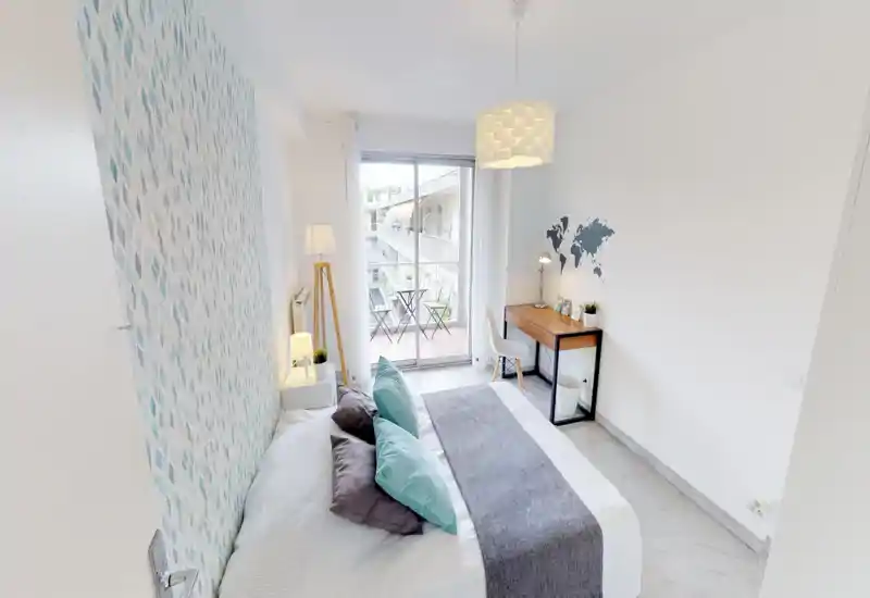 Chambre lumineuse et moderne avec un lit double habillé de linge neutre, coussins décoratifs et une tête de lit texturée ; bureau et balcon avec sièges offrent un coin travail et détente.