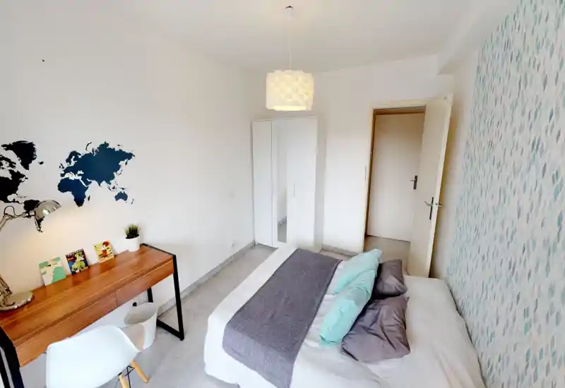 Chambre moderne et lumineuse avec un lit double habillé de linge neutre, coussins décoratifs et un mur d'accent bleu; comprend un bureau en bois et une armoire blanche.