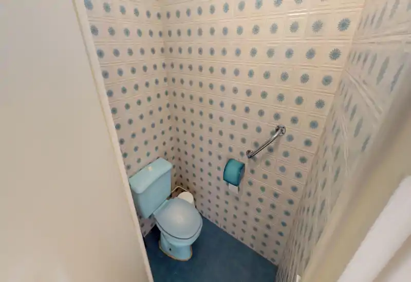 Petite salle de bains avec WC bleu et carreaux muraux vintage à motif ; espace compact avec barre d'appui et porte-papier toilette bleu assorti.