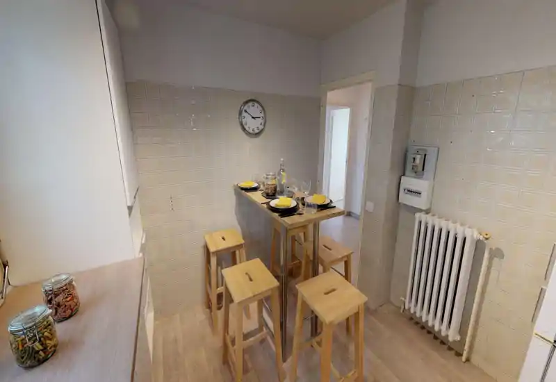 Cuisine compacte avec une table haute en bois et quatre tabourets, murs carrelés et bocaux sur le plan de travail — bien éclairée et aménagée pour des repas informels.