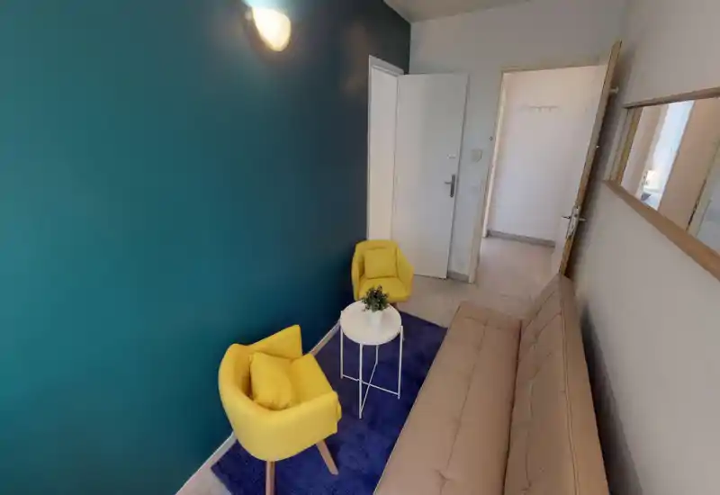 Coin salon compact avec un mur accent teal, deux fauteuils jaunes, une petite table ronde sur un tapis bleu et un canapé beige — lumineux, stylé et bien mis en scène pour un espace de détente.