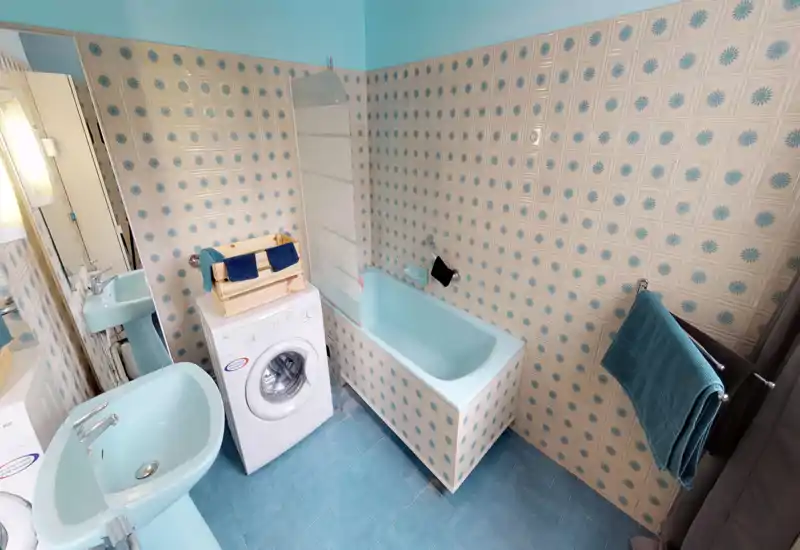 Salle de bain rétro et lumineuse avec carrelage bleu, baignoire et lavabos assortis, et une machine à laver à chargement frontal — espace propre et fonctionnel.