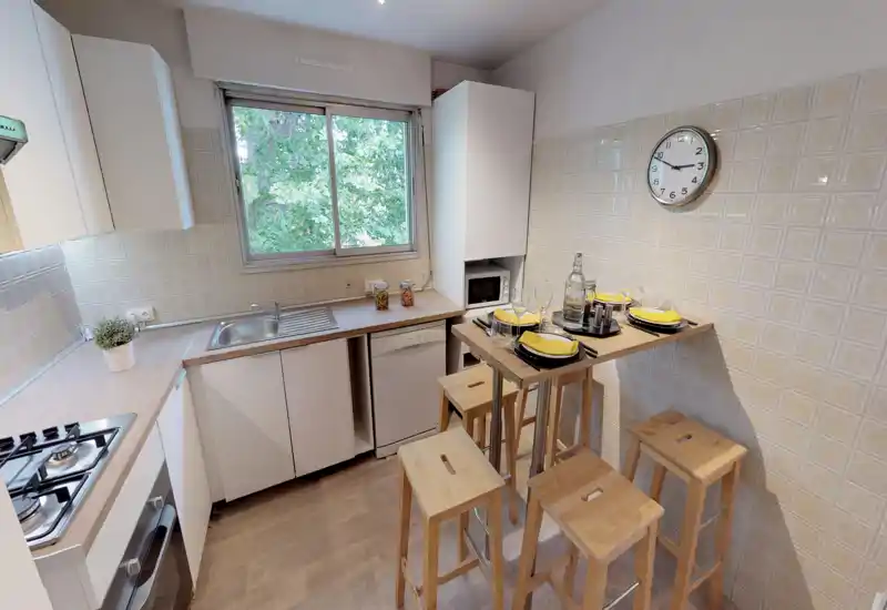 Cuisine compacte et bien équipée avec évier, plaque à gaz, four encastré et un comptoir haut avec tabourets en bois — fenêtre lumineuse et table dressée pour quatre.