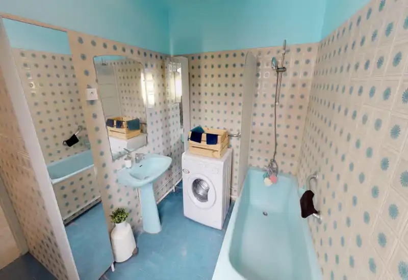 Salle de bain au style rétro avec baignoire et lavabo turquoise, carreaux à motif et machine à laver ; espace lumineux avec miroir mural et pommeau de douche.