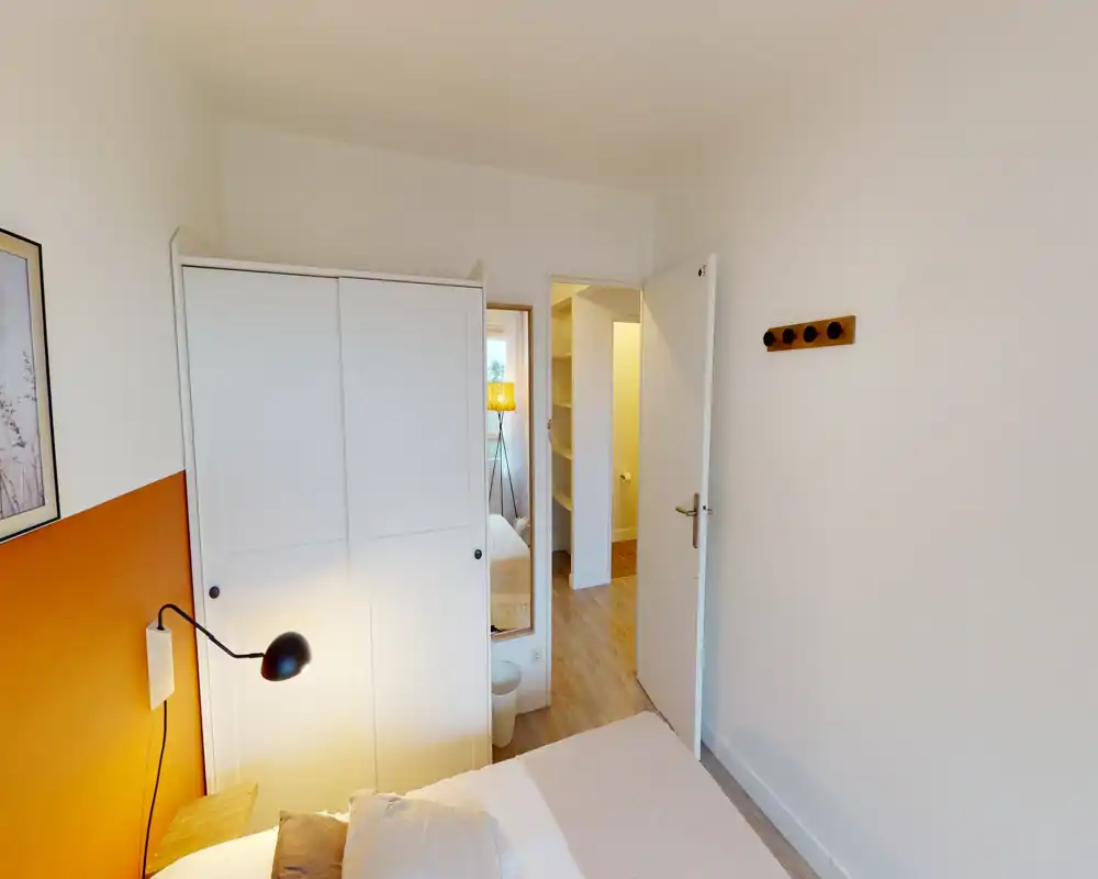 Camera da letto luminosa e accogliente con armadio bianco, lampada da comodino e parete d'accento calda. Il letto è il punto focale e la stanza mostra pavimento in legno e arredi moderni e semplici.