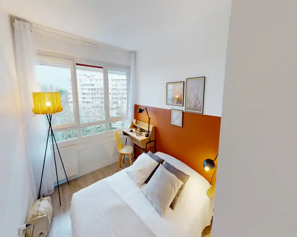 Camera da letto luminosa e accogliente con letto matrimoniale, parete testiera arancione, lampade da comodino e una scrivania piccola vicino alla finestra. Ampie finestre garantiscono molta luce naturale e l’ambiente è arredato con biancheria neutra e quadri incorniciati.