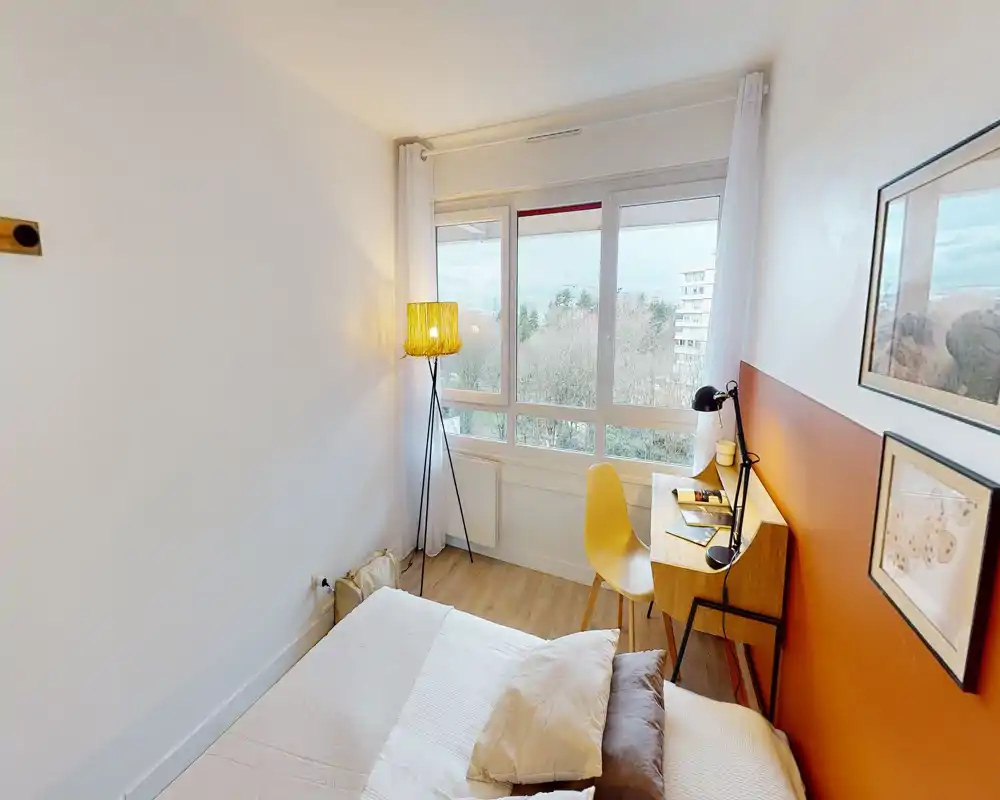 Camera da letto luminosa e compatta con letto confortevole, una piccola scrivania in legno e una sedia gialla davanti a una grande finestra con vista — accogliente, ben illuminata e arredata con gusto per una persona.