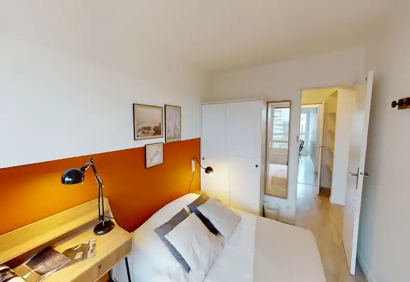 Camera da letto calda e luminosa con parete d'accento arancione, letto matrimoniale con cuscini sovrapposti, scrittoio in legno e lampada da lettura. L'ambiente è accogliente e moderno, con armadio a muro e luce naturale dalla zona adiacente.