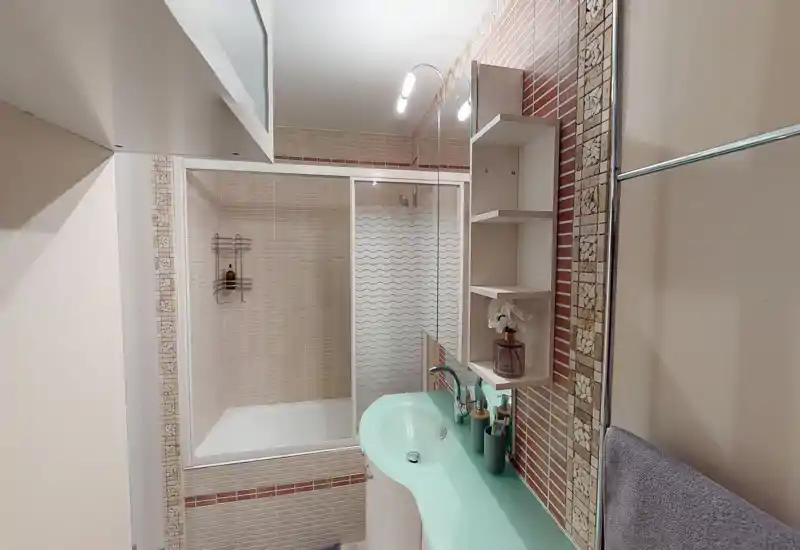 Salle de bains moderne et propre avec baignoire et paroi de douche en verre, plan vasque courbé vert menthe, étagères murales et carrelage aux tons neutres.