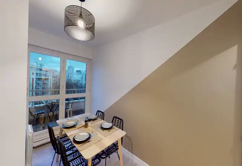 Coin repas moderne et lumineux pour quatre personnes, avec grande fenêtre et vue sur le balcon. Table compacte en bois, chaises à motif et suspension pour une ambiance chaleureuse et stylée.