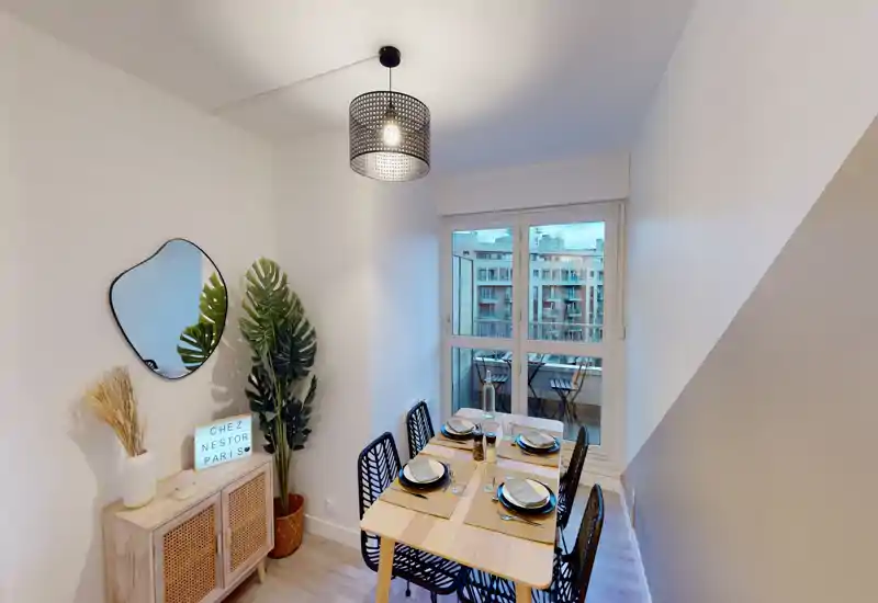 Coin repas lumineux et soigné avec une table en bois pour quatre, buffet décoratif, miroir et plantes ; vue urbaine moderne depuis la fenêtre.