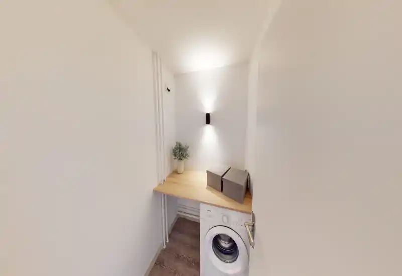 Niche buanderie/utility compacte avec machine à laver frontal, étagère en bois au-dessus pour le rangement et applique murale diffusant une lumière douce — sobre et optimisée.