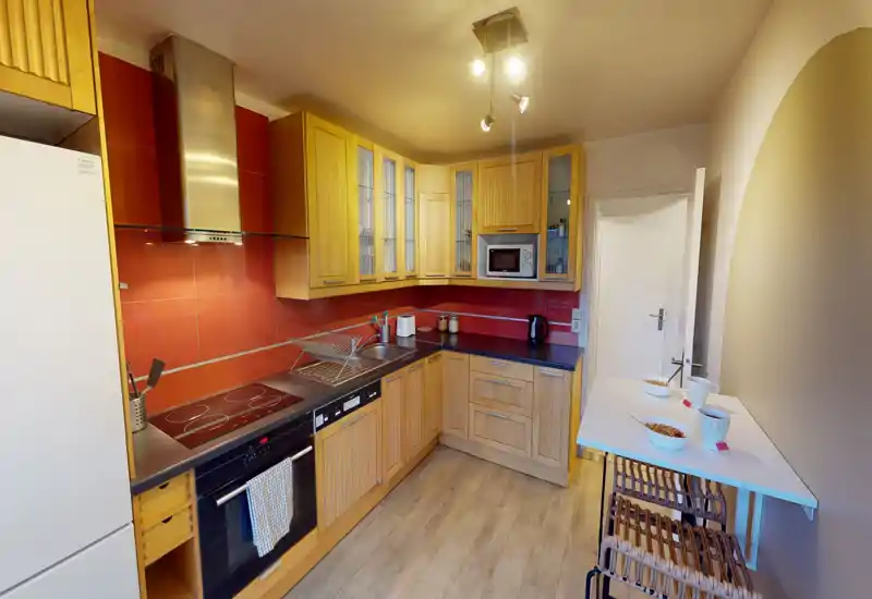 Cuisine compacte et bien équipée avec placards en bois, plaque vitrocéramique, four et micro-ondes encastrés, et un petit comptoir — finitions chaudes et lumineuses, agencement pratique adapté aux locations courte durée.