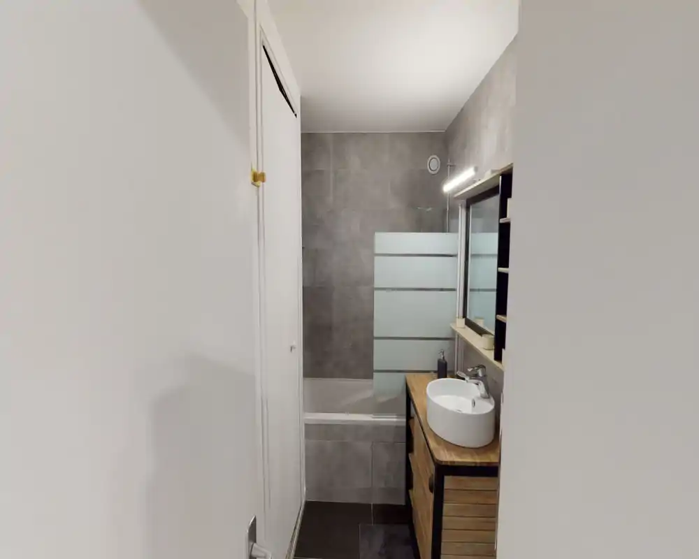 Modern compact bathroom with a bathtub, tiled wall, wooden vanity and round countertop sink; mirror and strip light above the sink.